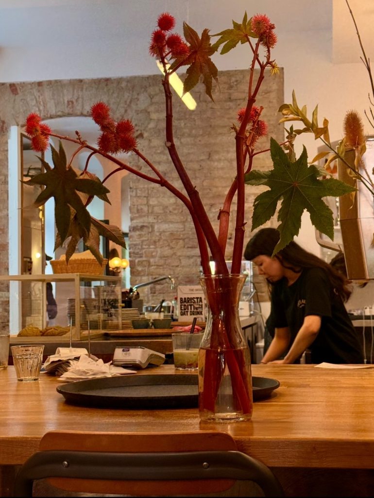 Deep red branches arranged in a glass vase on a communal wooden table inside a contemporary Japanese café while a barista works in the background.